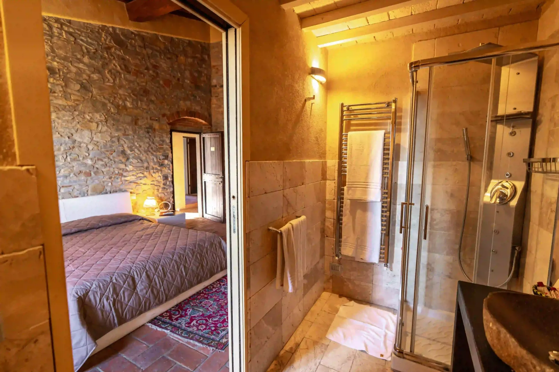 Vista da un bagno moderno con box doccia in vetro su una camera da letto rustica adiacente con pareti in pietra e un letto.