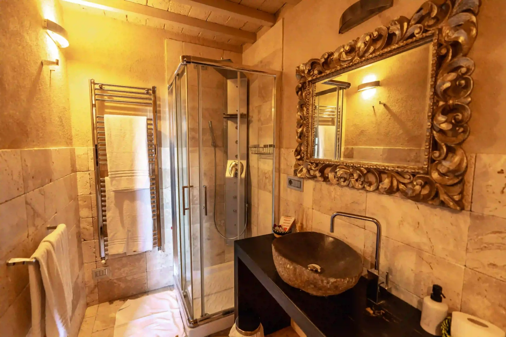 Un bagno dai toni caldi, con pareti in pietra, dotato di doccia in vetro, lavabo in pietra su mobiletto nero e un elaborato specchio dorato.