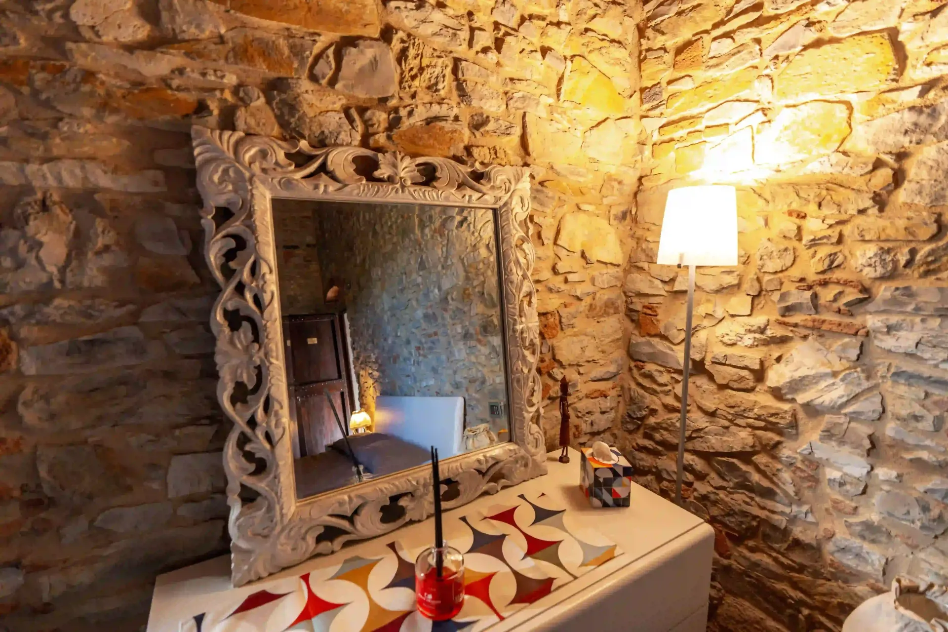 Uno specchio decorato con una cornice bianca è appoggiato su una toeletta decorata, di fronte a una parete in pietra rustica, con una lampada da terra accesa nelle vicinanze.