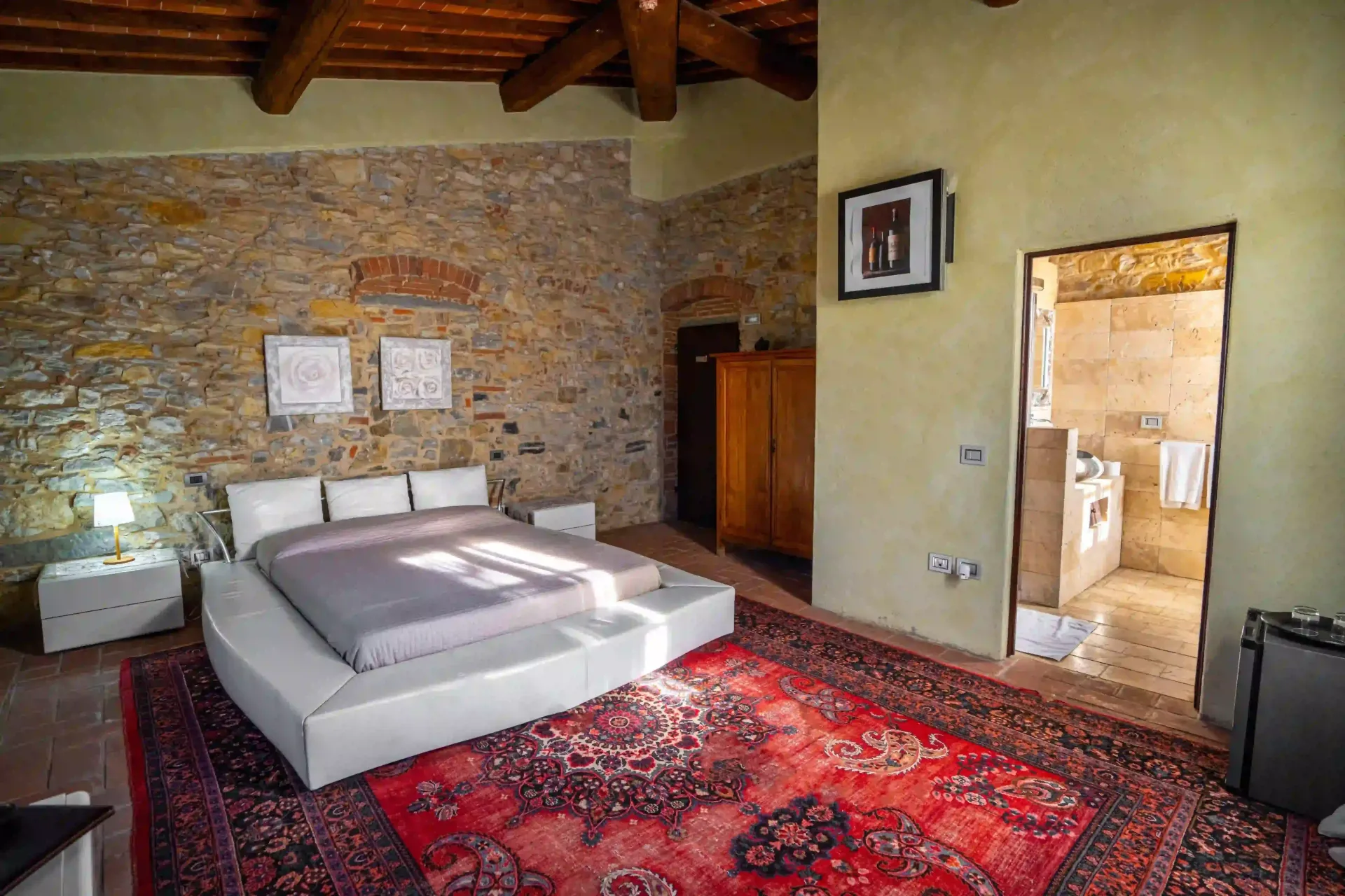 Una camera da letto con un letto basso bianco, un grande tappeto rosso decorato, pareti in pietra rustica, travi a vista sul soffitto e un bagno a vista.