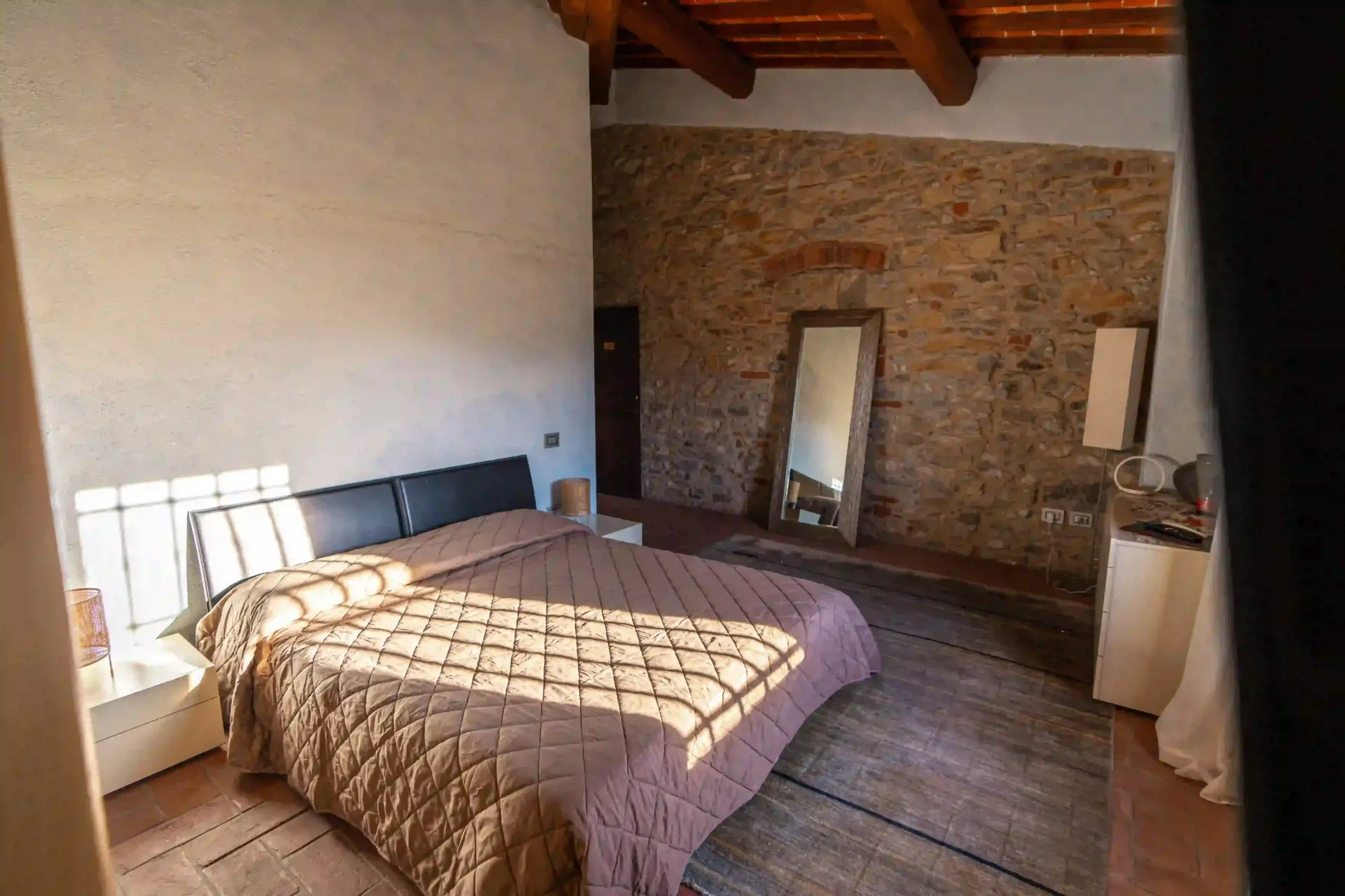Una camera da letto con un letto trapuntato marrone, una parete in pietra a vista, travi in legno a vista sul soffitto e uno specchio a figura intera.