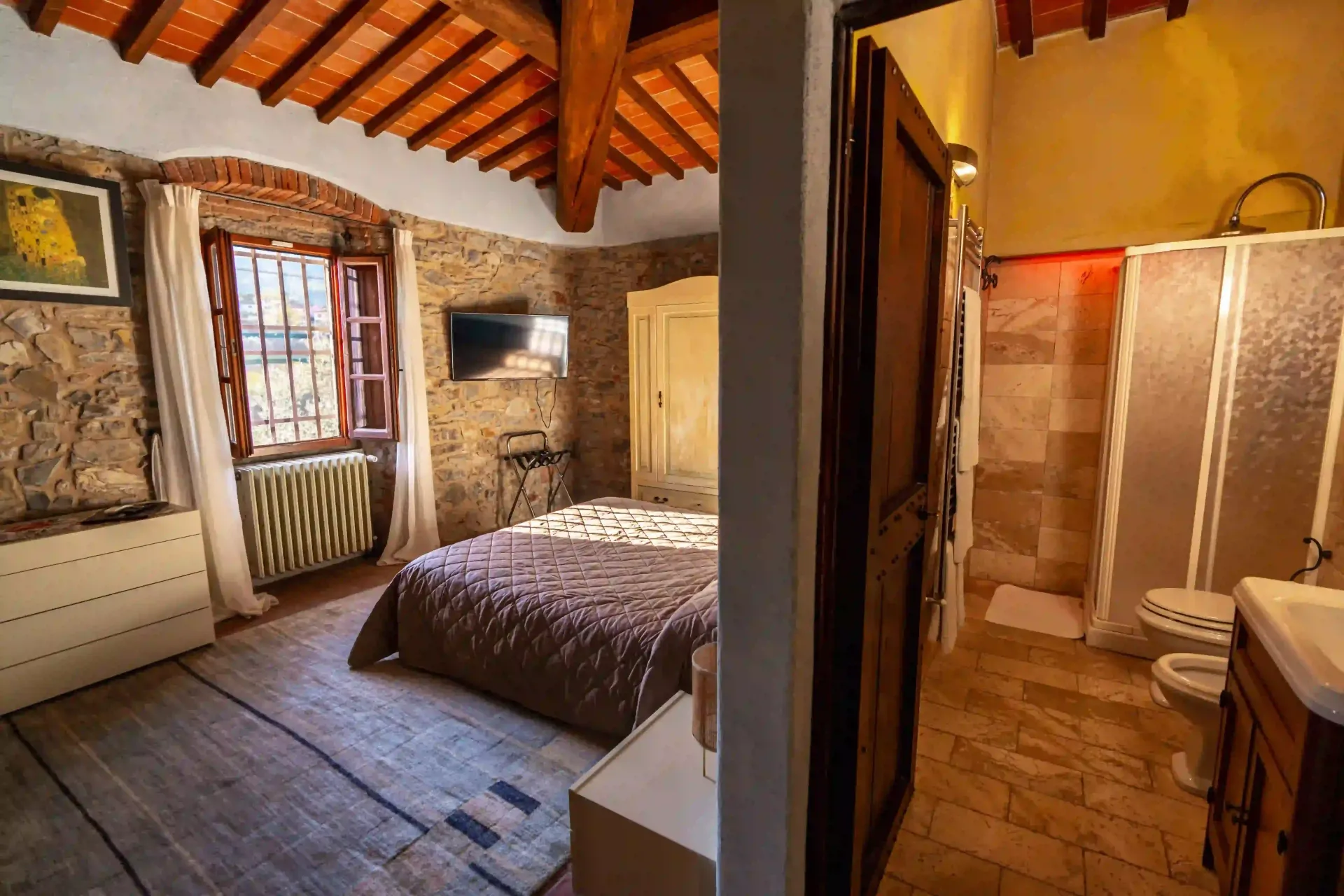 Una camera da letto rustica con pareti in pietra, travi in legno a vista e un letto, adiacente a un bagno a pianta aperta con pavimento in pietra.
