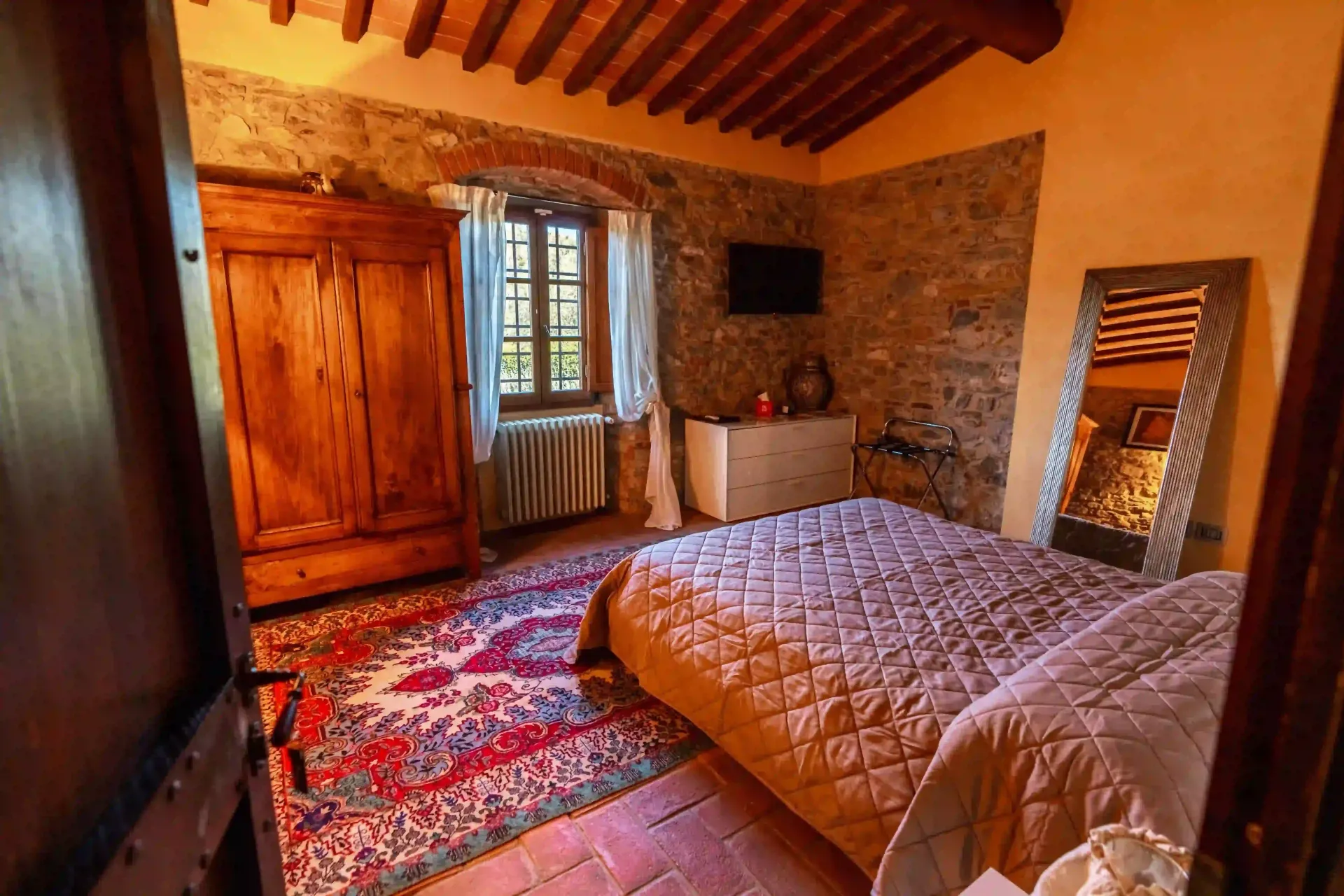 Una camera da letto accogliente e rustica con pareti in pietra, travi a vista sul soffitto, un letto trapuntato, un grande armadio in legno e un tappeto rosso.