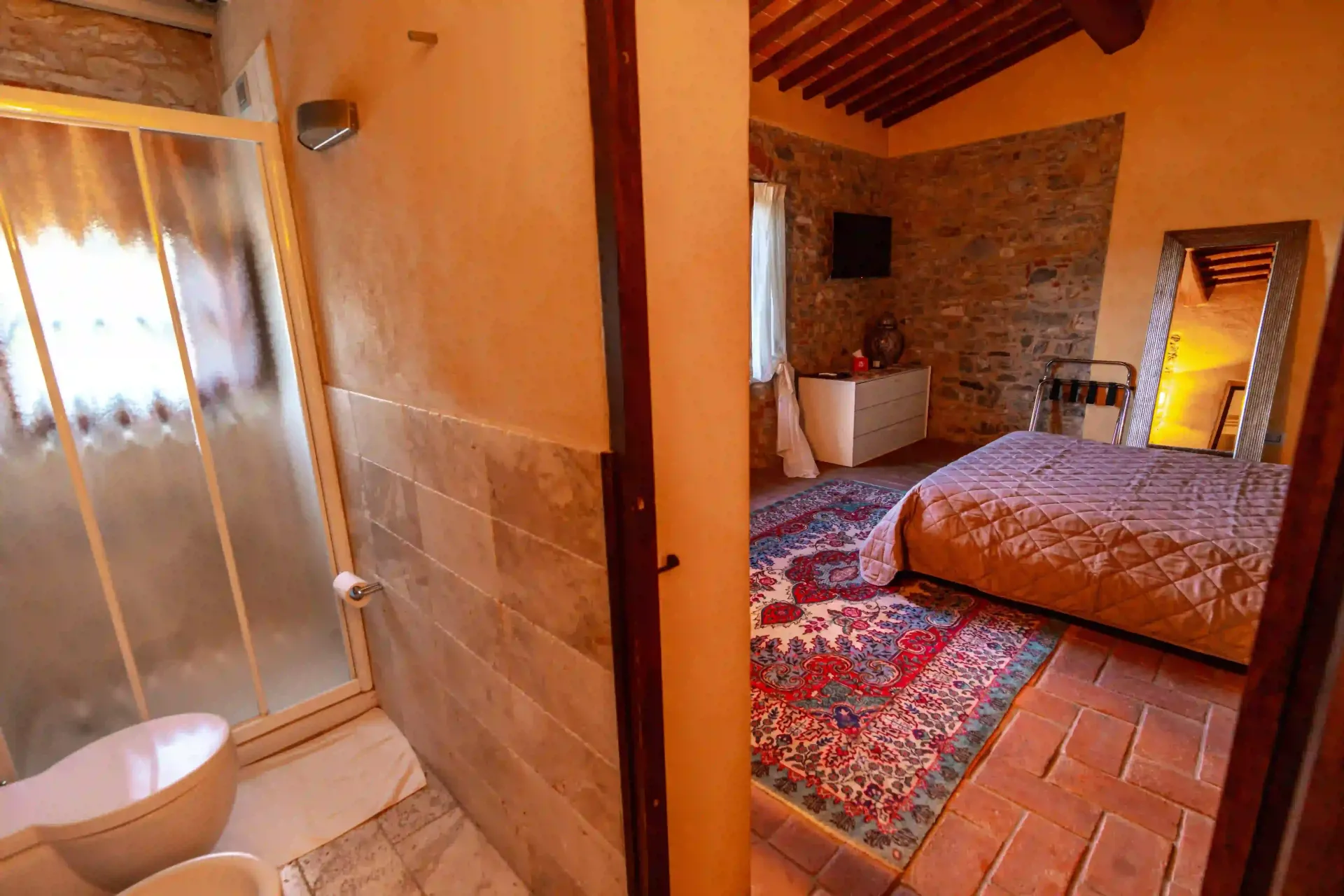 Una vista da un bagno piastrellato che si affaccia su una camera da letto rustica con un tappeto a motivi geometrici, un letto e uno specchio decorativo.