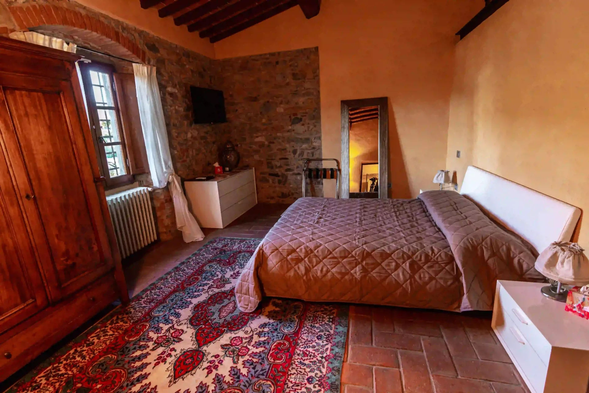 Una camera da letto rustica con letto matrimoniale, tappeto decorato, armadio in legno, parete in pietra e pavimento in cotto sotto un soffitto con travi a vista.