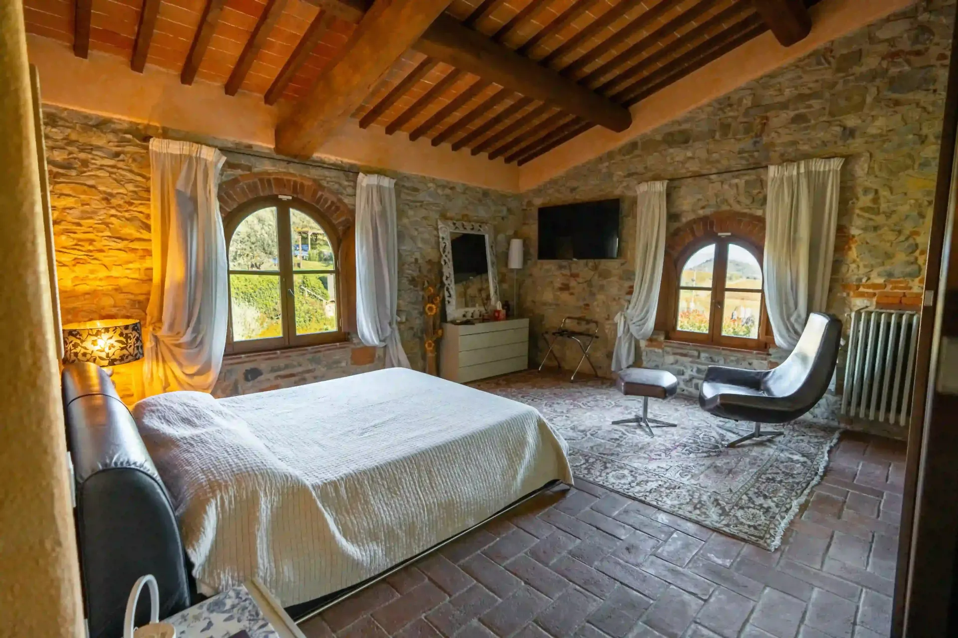 Una camera da letto rustica con pareti in pietra, pavimento in cotto, un letto matrimoniale e una poltrona in pelle accanto alla finestra.