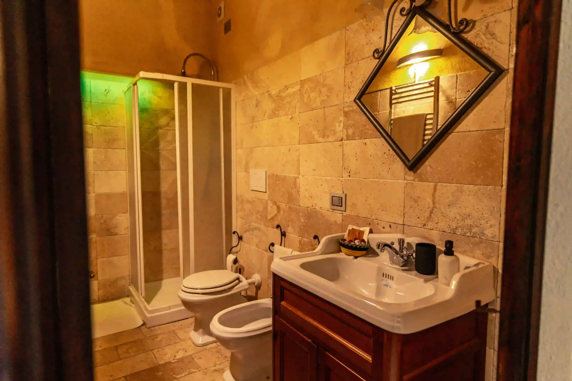 Un bagno rustico con pareti rivestite in travertino, box doccia, WC, bidet e mobiletto in legno con specchio.