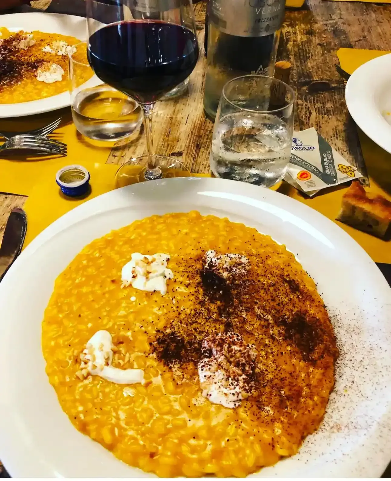 Su un tavolo, due piatti di risotto giallo con panna bianca e spezie scure, accompagnati da bicchieri di vino rosso e acqua.