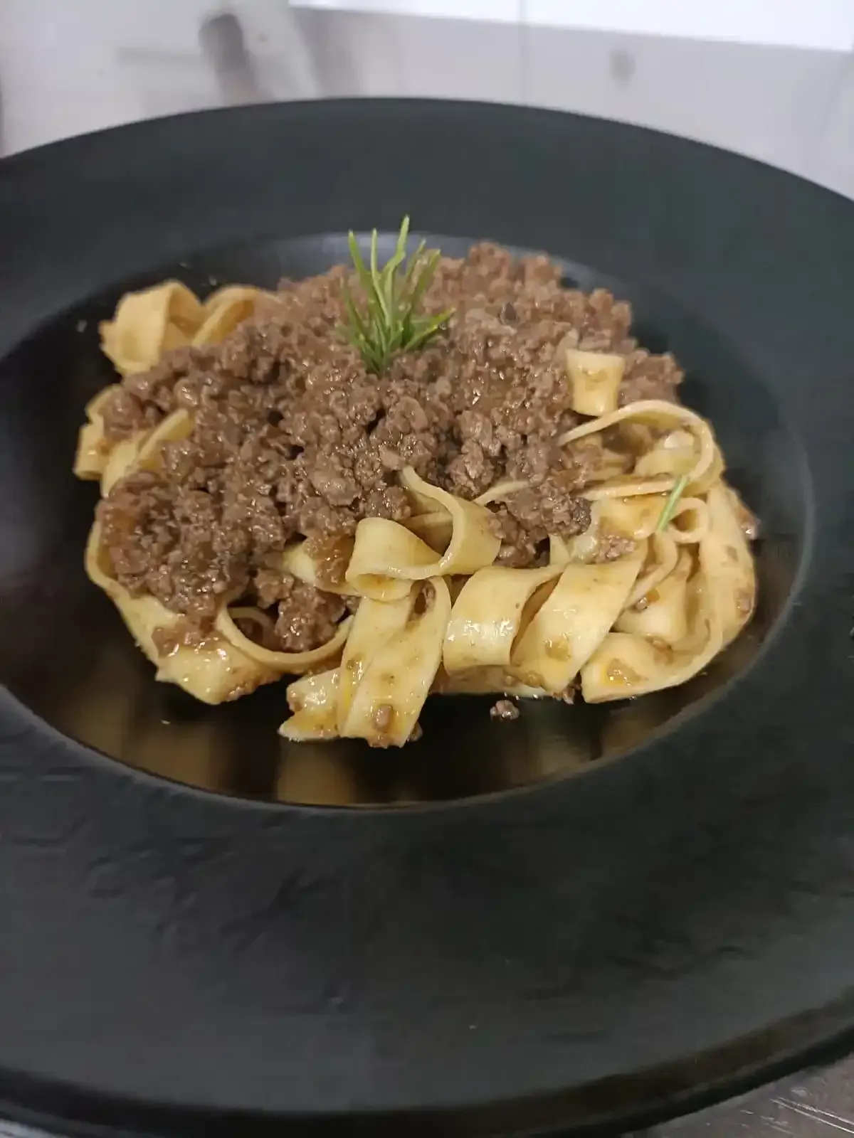 Un mucchio di pasta con un ricco ragù di carne, guarnito con un rametto di erbe aromatiche, servito in un'ampia ciotola nera.