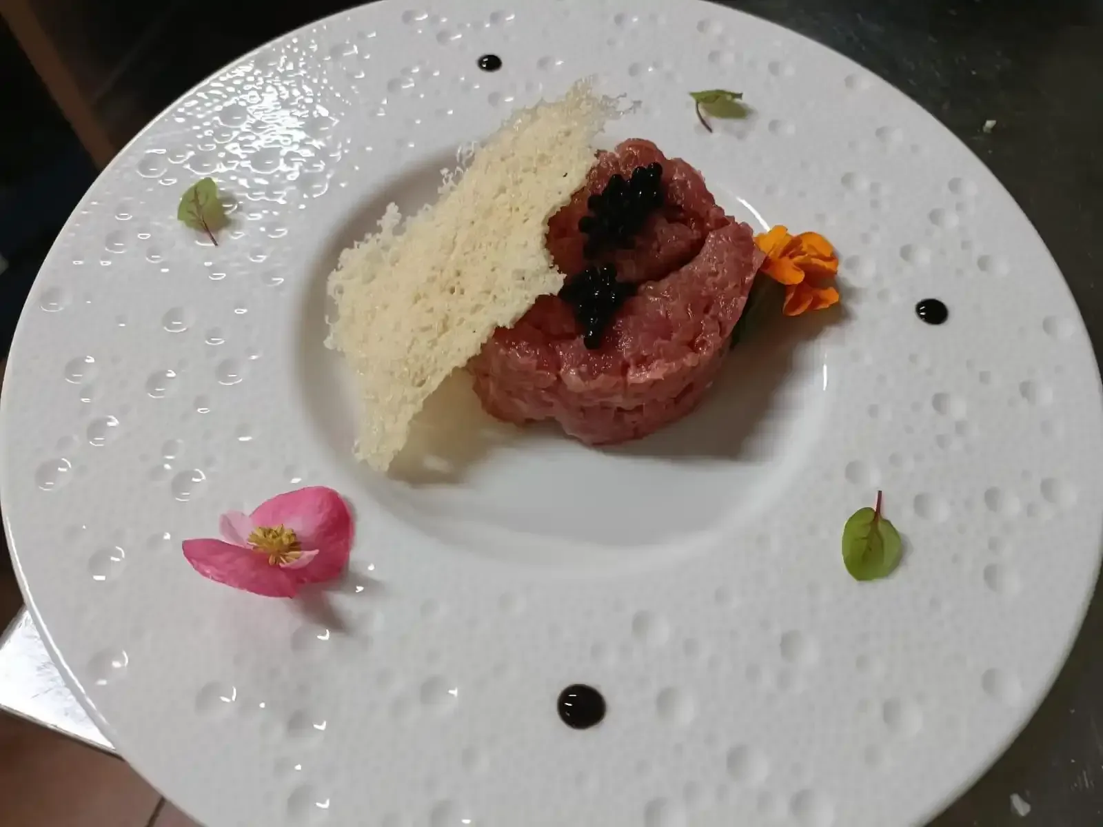 Tartare di manzo con caviale nero e una cialda di parmigiano, guarnita con fiori su un piatto bianco dalla superficie ruvida.