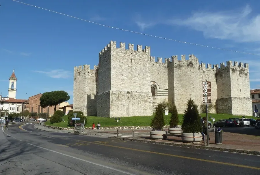 Un castello medievale in pietra chiara, con mura merlate, sorge a lato di una strada sotto un cielo azzurro brillante.