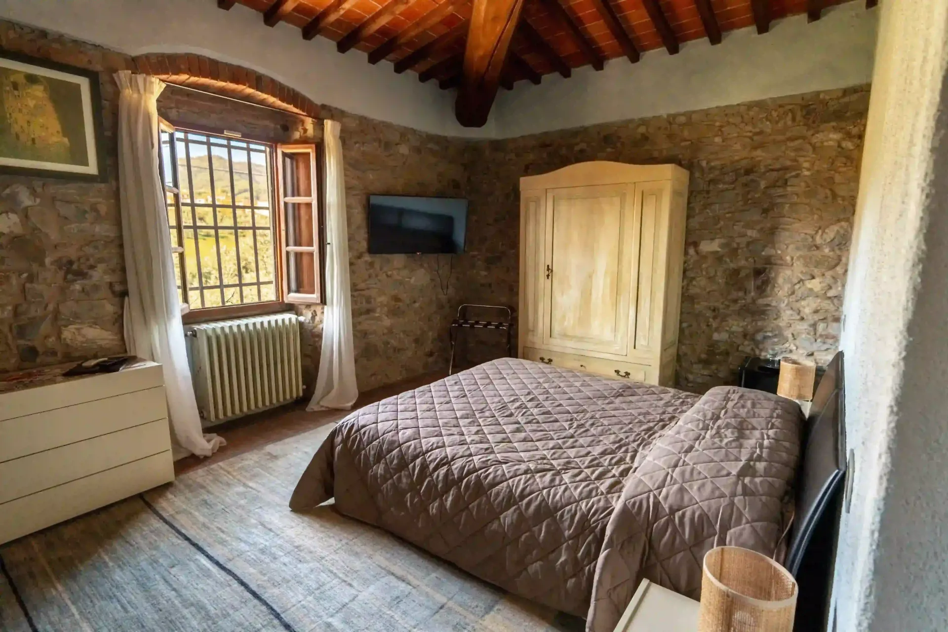 Una camera da letto accogliente con pareti in pietra, soffitto in legno, letto con copriletto trapuntato marrone, armadio e finestra con vista.