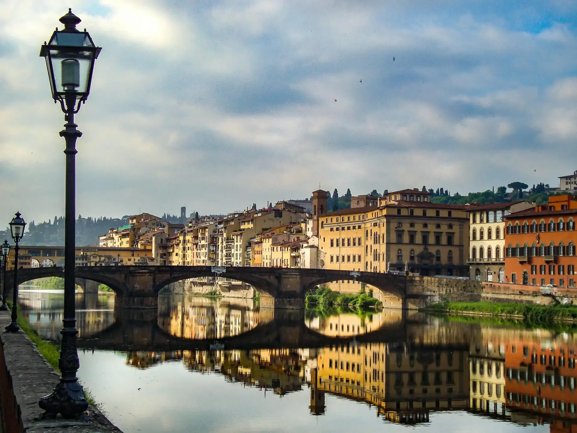 In primo piano, un lampione in ferro battuto riccamente decorato si affaccia su un ponte di pietra che attraversa un fiume, con sullo sfondo edifici storici di Firenze.