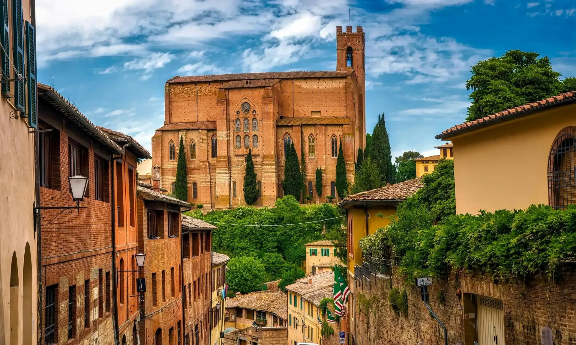 Una strada stretta e soleggiata, fiancheggiata da storici edifici in mattoni, conduce a una grande e riccamente decorata chiesa in mattoni sotto un cielo azzurro.
