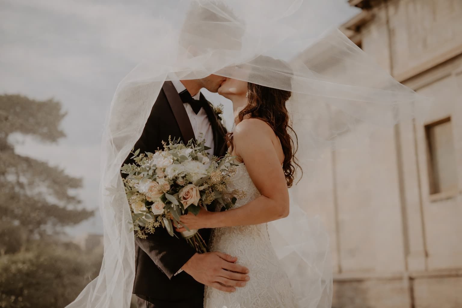 Gli sposi si baciano sotto un velo nuziale fluttuante, con la sposa che tiene in mano un bouquet di fiori.