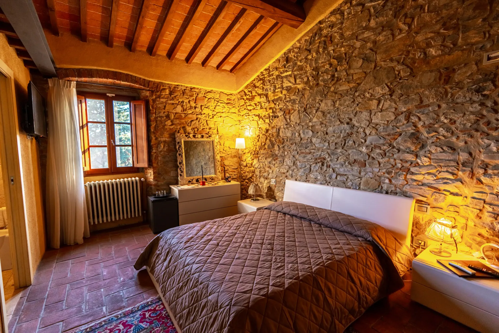 Una camera da letto rustica con pareti in pietra a vista, soffitto con travi in legno, una trapunta marrone sul letto e una finestra con tende.