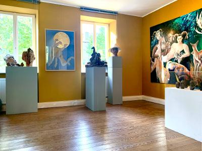 Kunstgalerie mit farbenfrohen Gemälden und Skulpturen in einem hellen Raum mit Holzboden.