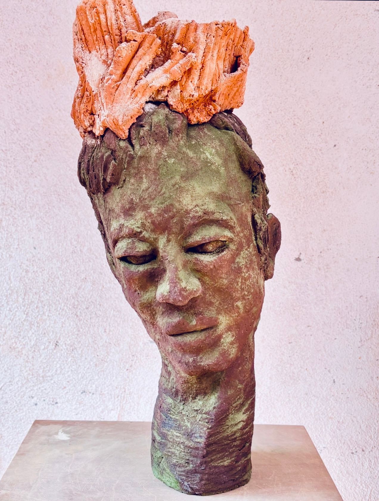 Abstrakte Skulptur eines Kopfes mit geschlossenen Augen und orangefarbenem, strukturiertem Haar auf einem Sockel.