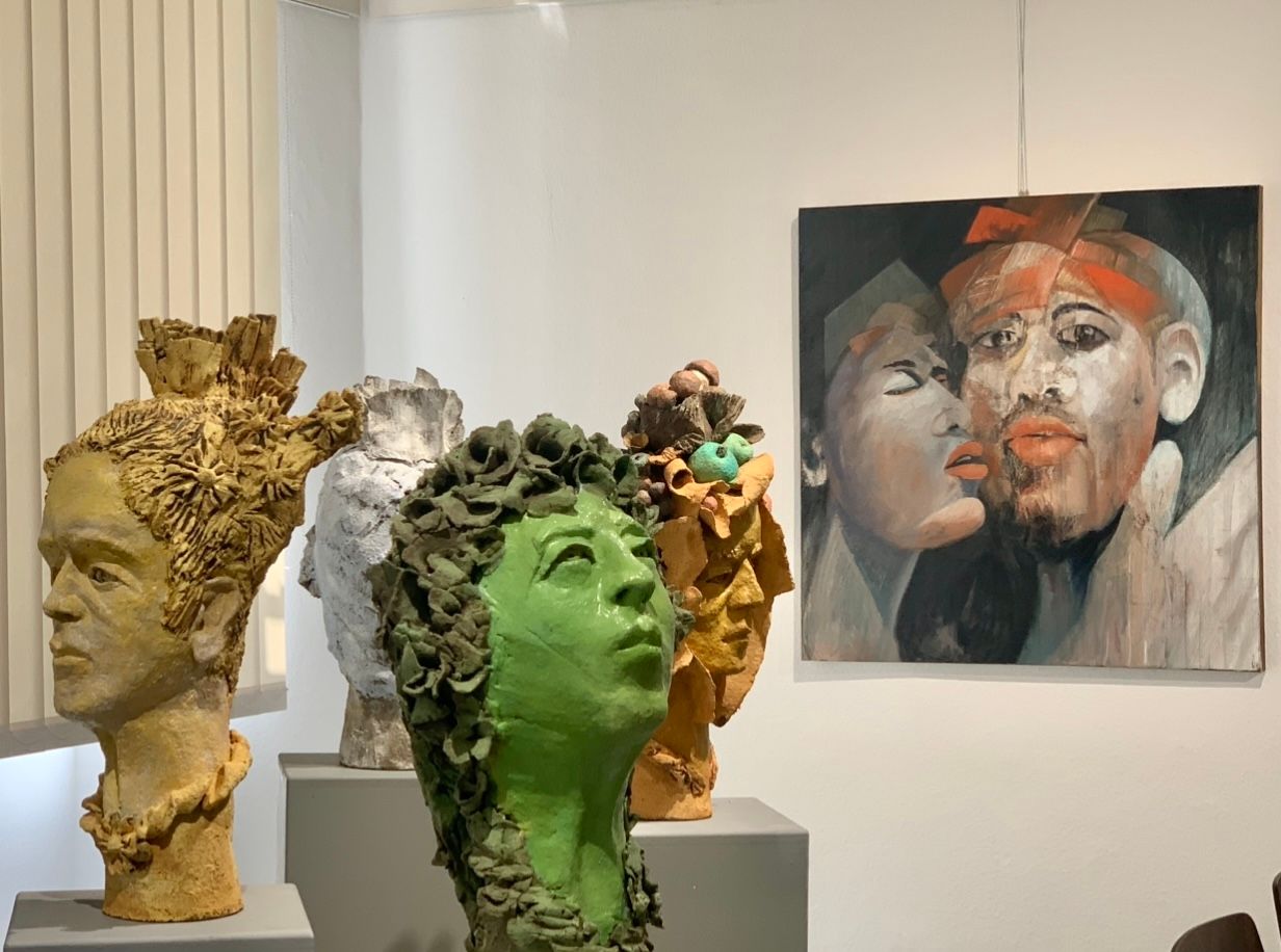In einer Kunstgalerie sind Skulpturenköpfe in Gold, Weiß und Grün neben einem gemalten Porträt an der Wand ausgestellt.