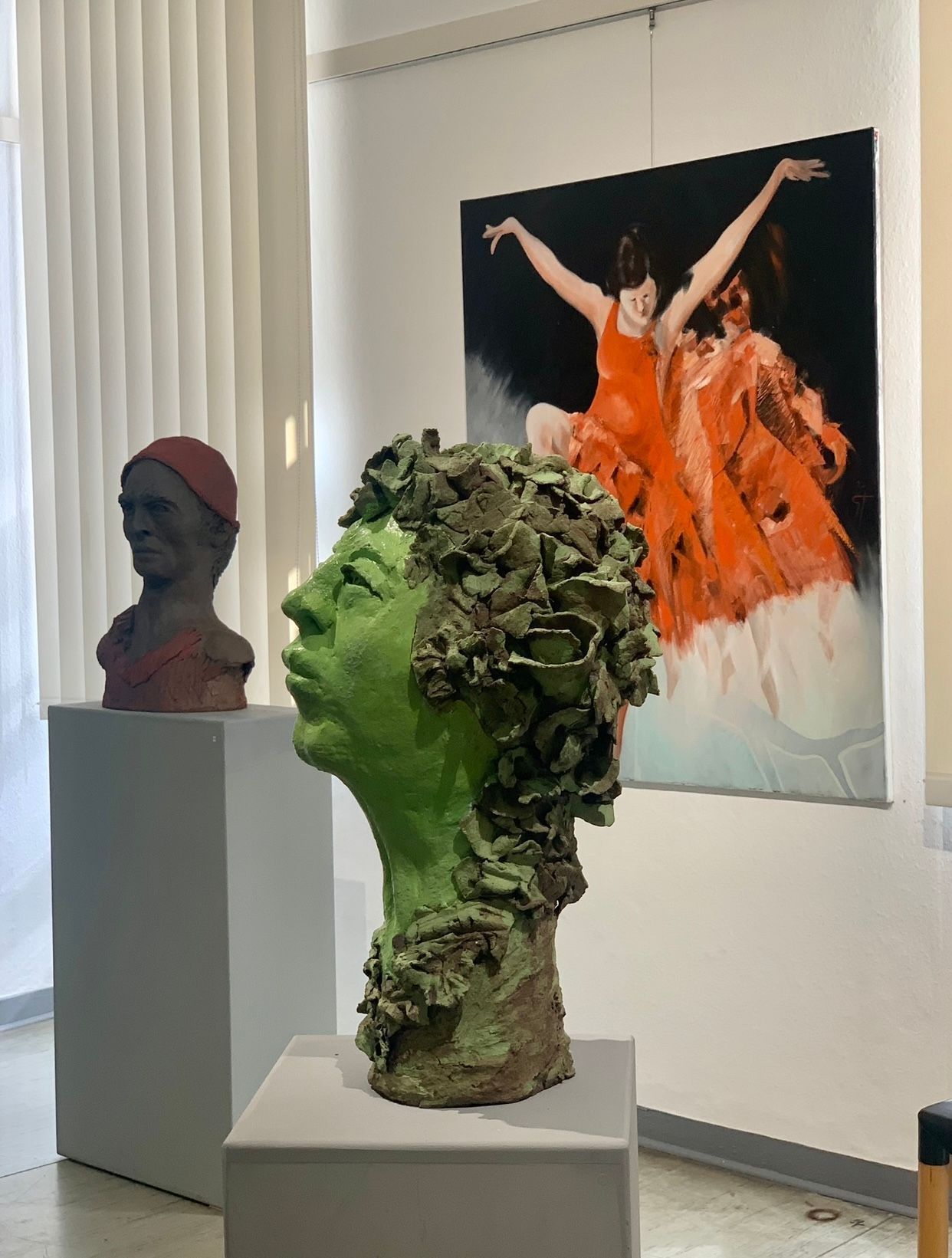 Galerieausstellung mit zwei Skulpturenköpfen auf Sockeln, im Hintergrund eine orangefarbene Figurenskulptur.