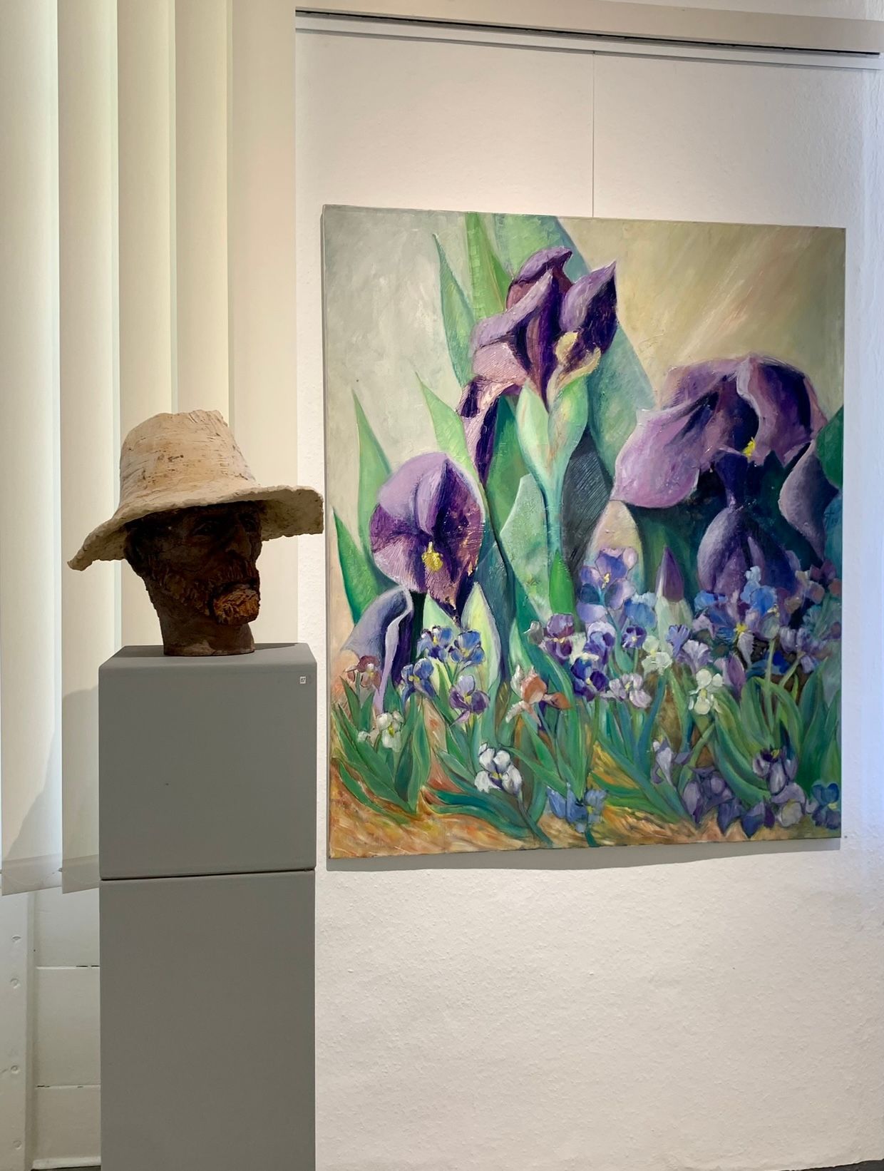 Galerieausstellung mit einer Skulptur mit Strohhut neben einem farbenfrohen Gemälde von violetten Schwertlilien.
