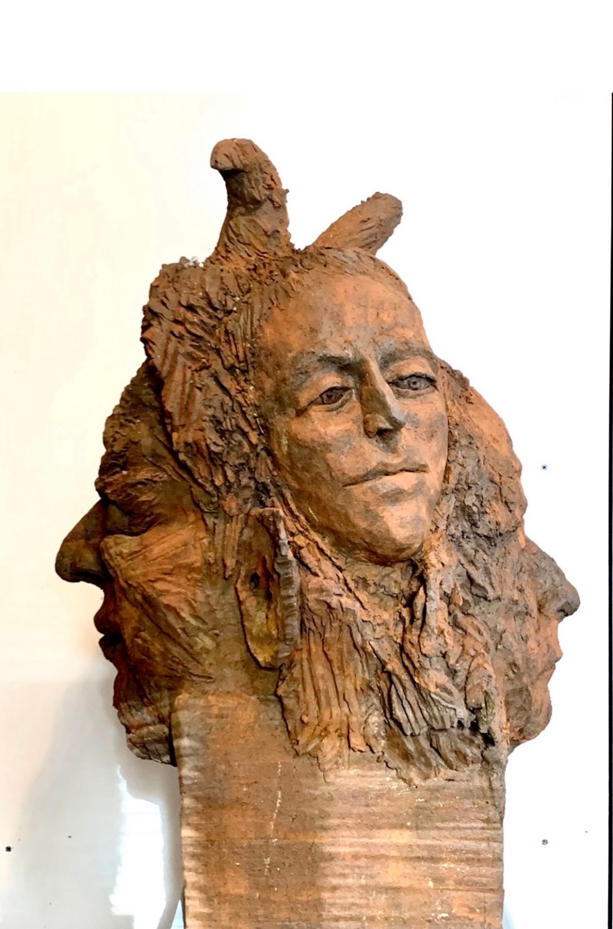 Holzskulptur eines menschlichen Gesichts mit strukturierten, haarähnlichen Schnitzereien und zwei nach oben gerichteten Ausbuchtungen vor einer weißen Wand