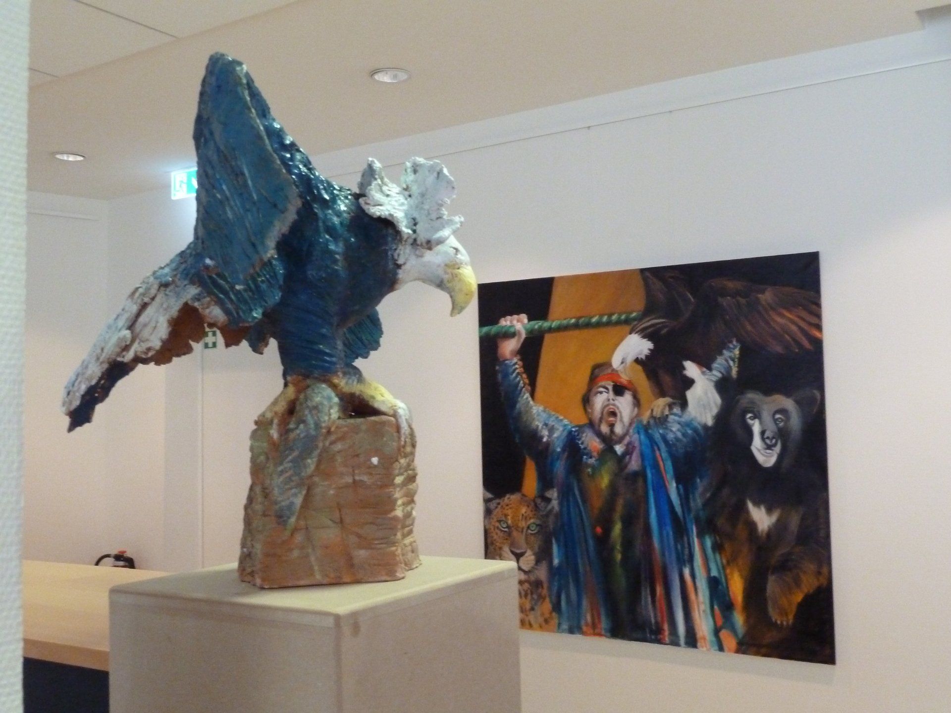 Abstrakte blaue Skulptur auf einem Sockel in einer Galerie, im Hintergrund ein farbenfrohes Gemälde.