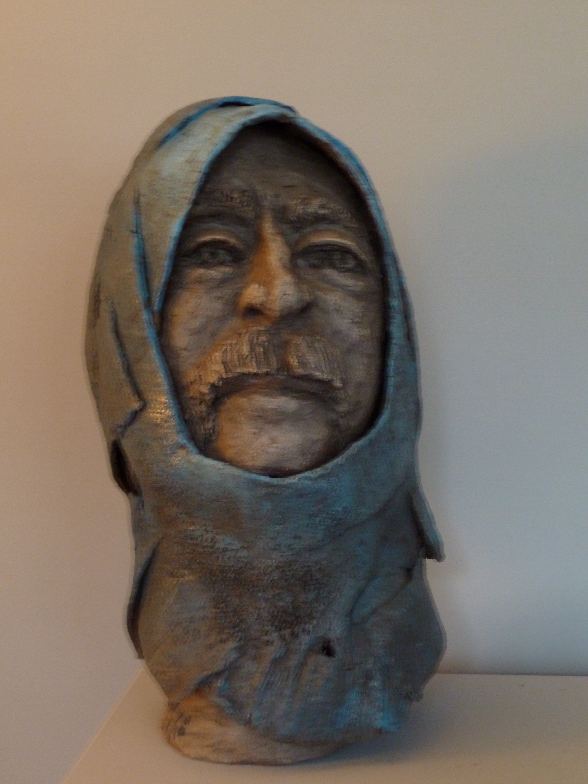 Skulptierte Büste eines Gesichts mit Kapuze in Graublautönen vor einem schlichten beigen Hintergrund
