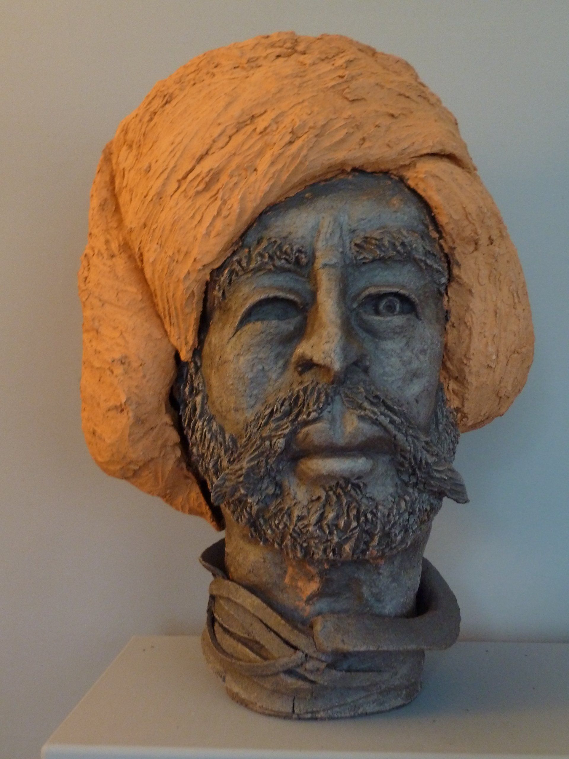 Tonbüste eines bärtigen Mannes mit orangefarbenem Turban auf einem Sockel vor schlichtem Hintergrund