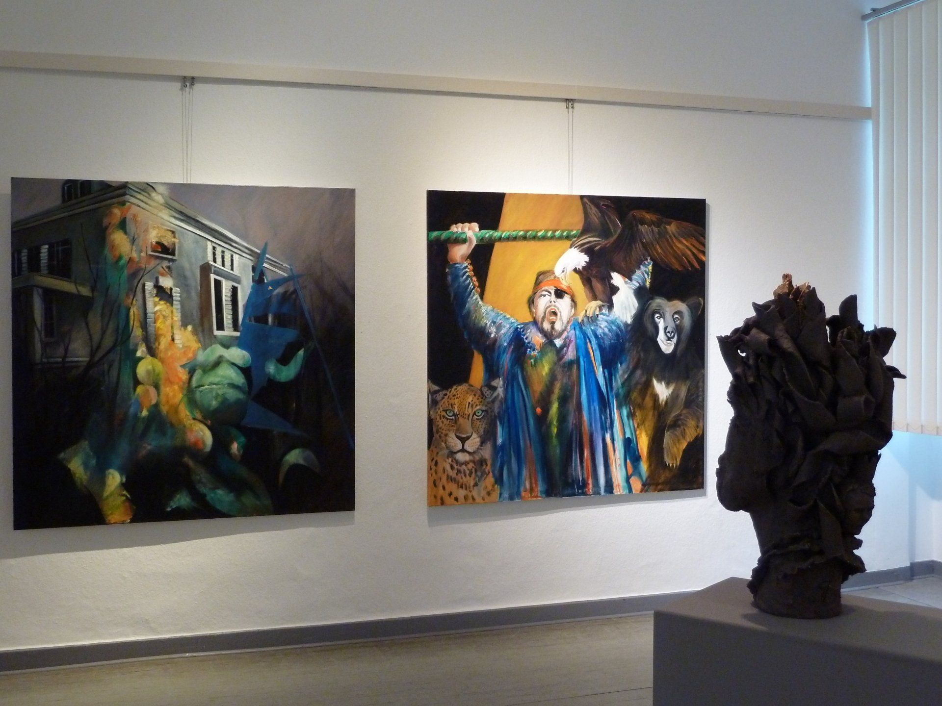 Wand in einer Kunstgalerie mit zwei farbenfrohen Gemälden und einer dunklen Skulptur auf der rechten Seite.