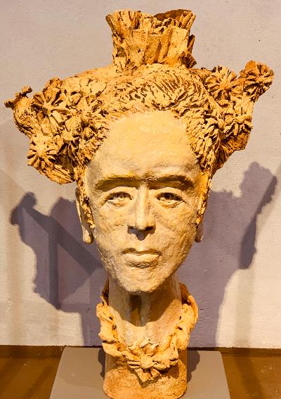Skulpturale Büste mit einem strukturierten, blumenartigen Kopfschmuck auf einem Sockel vor einer schlichten Wand.