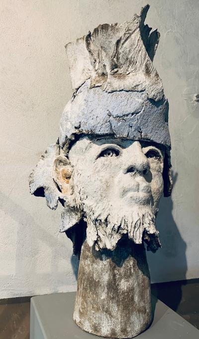 Verwitterte Steinskulptur eines menschlichen Kopfes mit hohem Kopfschmuck auf einem Sockel vor einer schlichten Wand