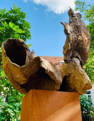 Abstrakte Holzskulptur auf einem Sockel im Freien, mit ausgehöhlten Ästen vor blauem Himmel und Bäumen.