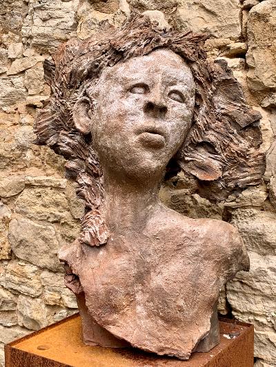 Steinskulptur einer Person mit lockigem Haar vor einer rauen Felswand, auf einem Holzsockel