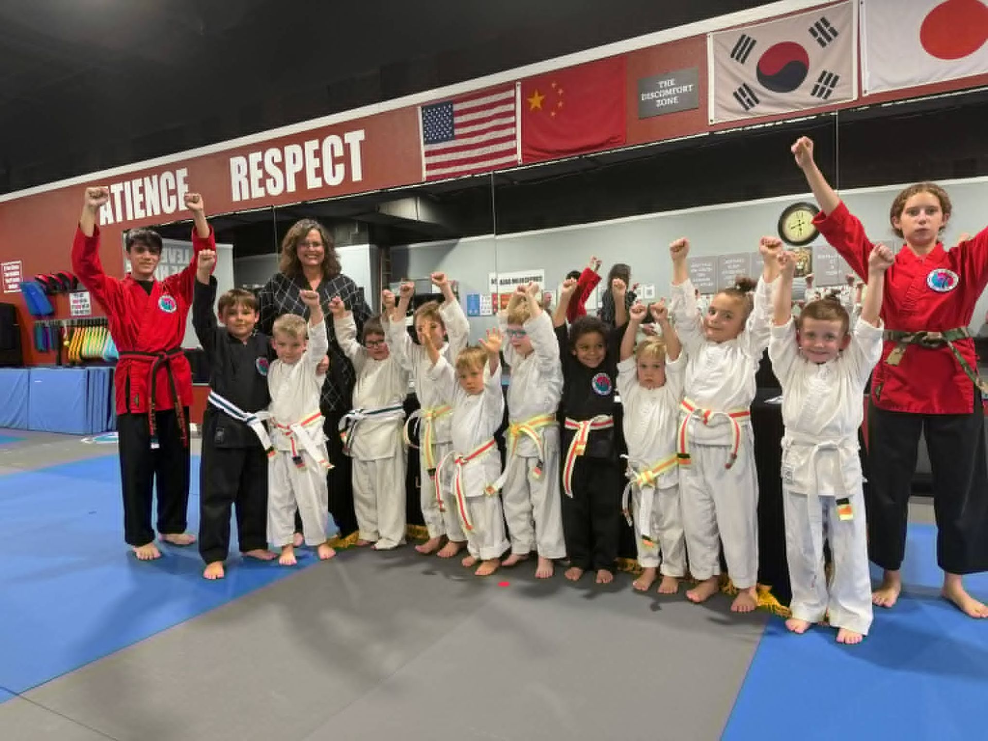 Children in white martial arts uniforms pose with instructors in a gym, holding medals and certificates.