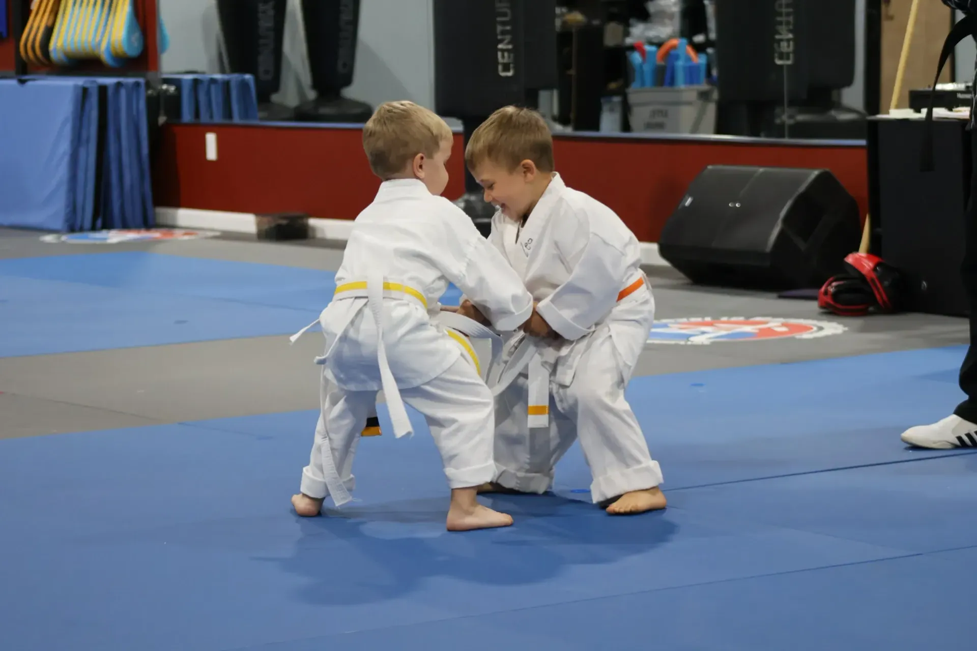 Two children in white gis sparring on a blue martial arts mat at an indoor tournament