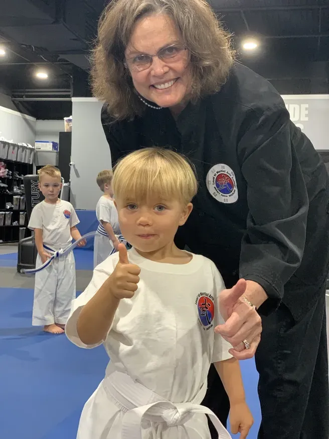 Smiling woman in black gi holds a child in a white gi giving a thumbs-up in a martial arts studio