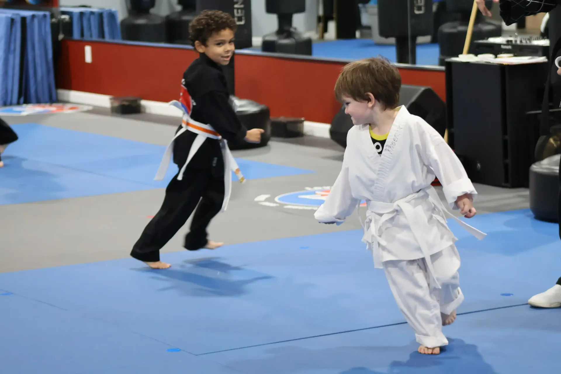 Two children in martial arts uniforms sparring on a blue mat in a gym