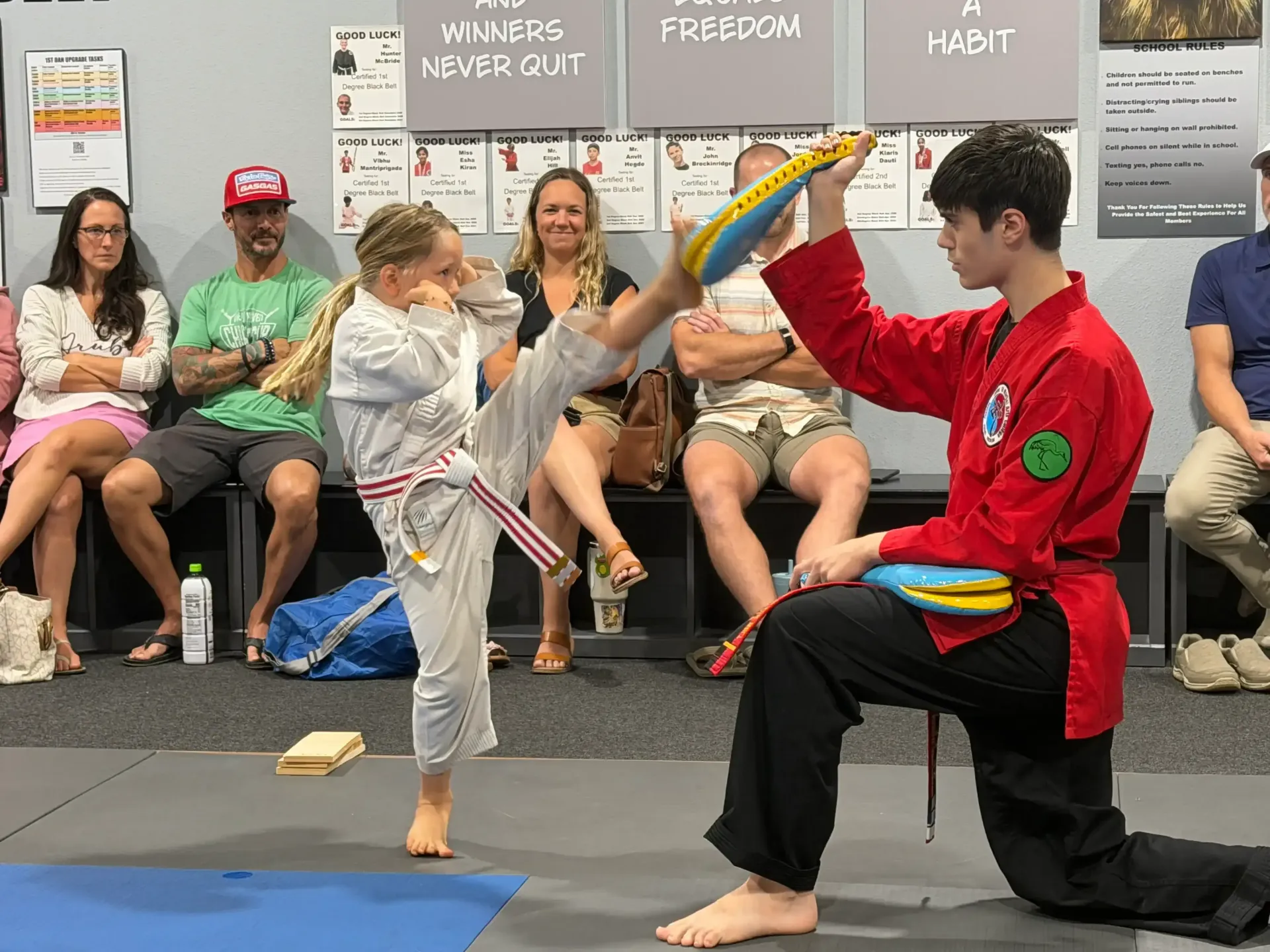 Two martial arts students spar with padded sticks while seated spectators watch indoors