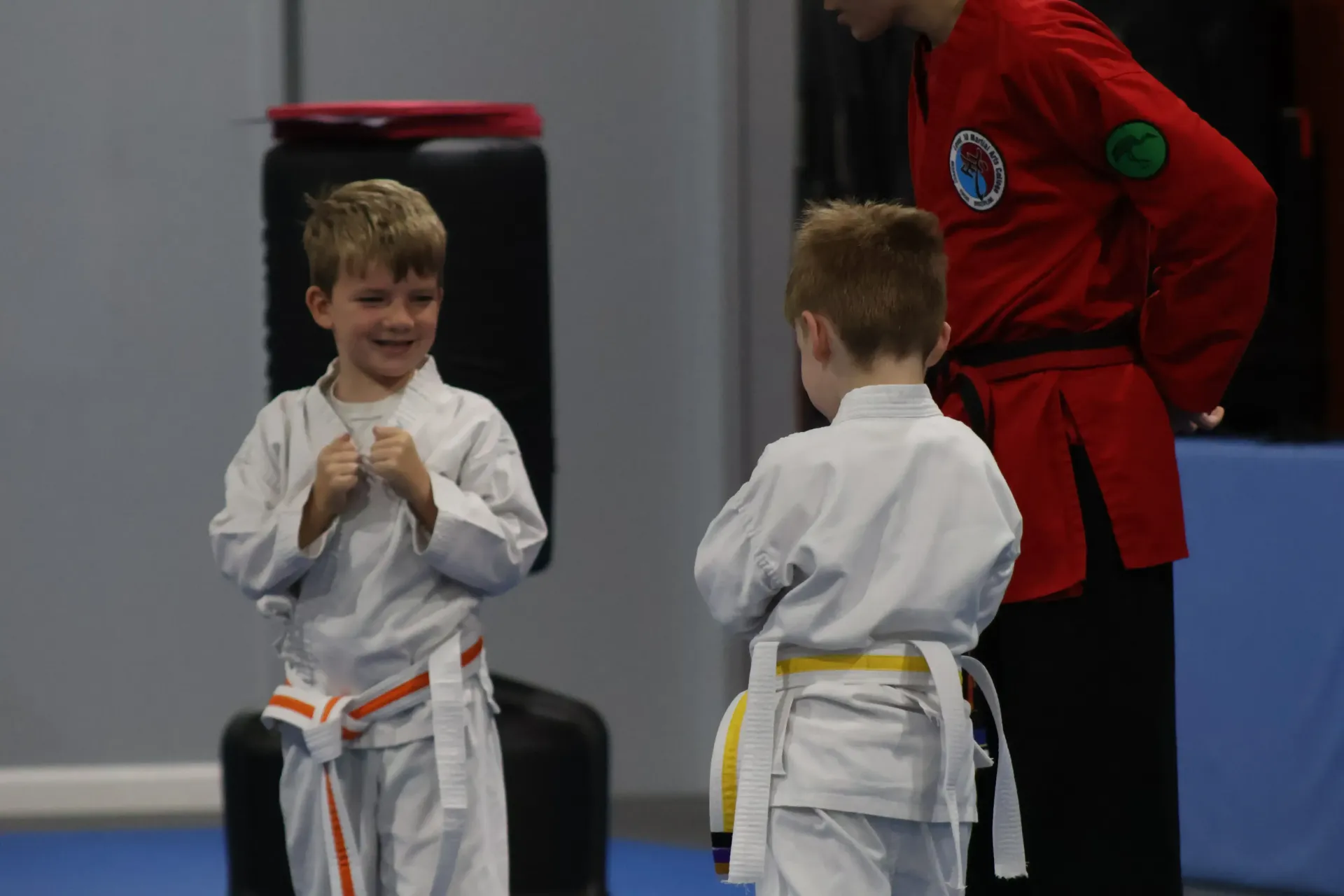 Two children in white martial arts uniforms stand on a mat, smiling and facing each other during practice.