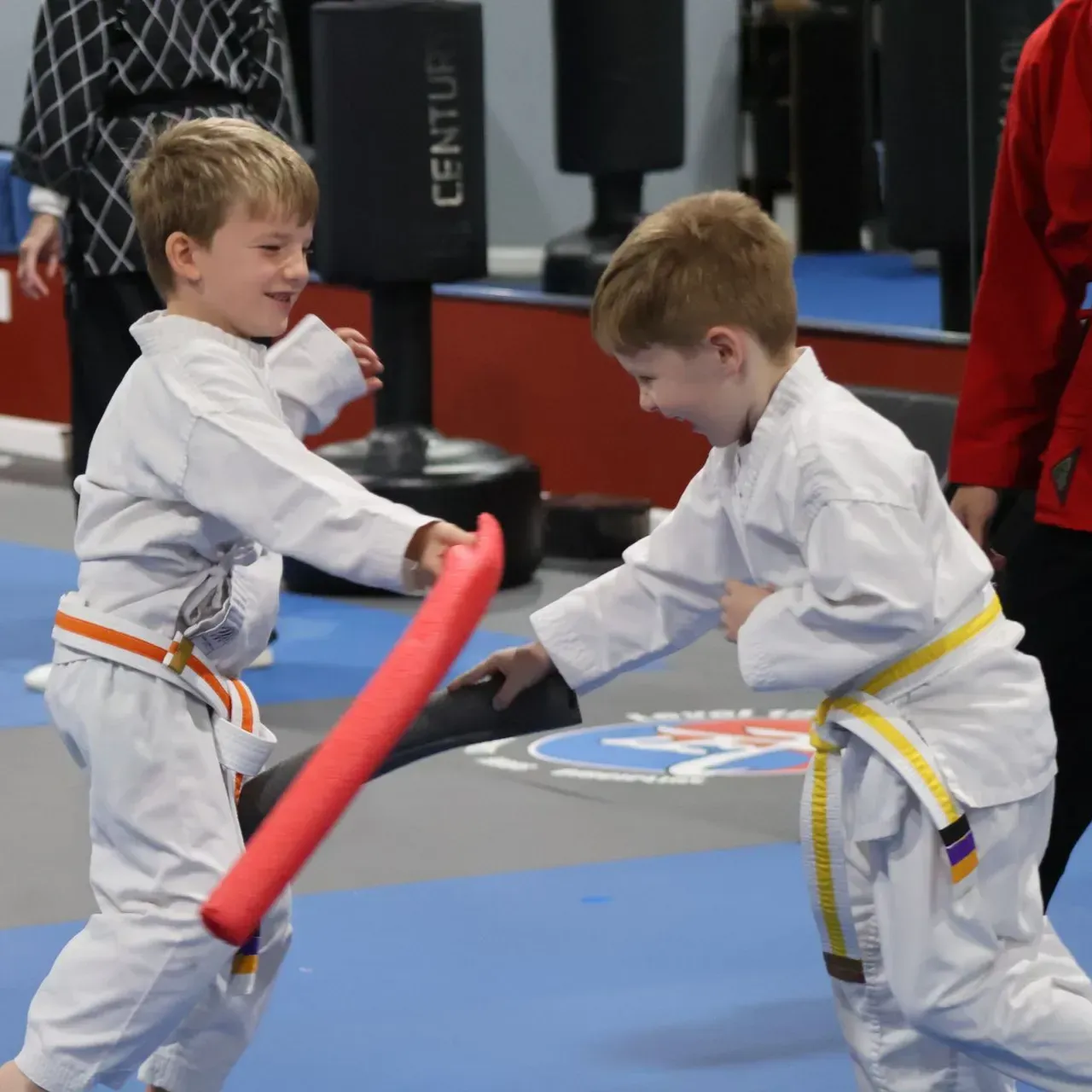 Two kids in white martial arts uniforms sparring with red foam swords in a gym.