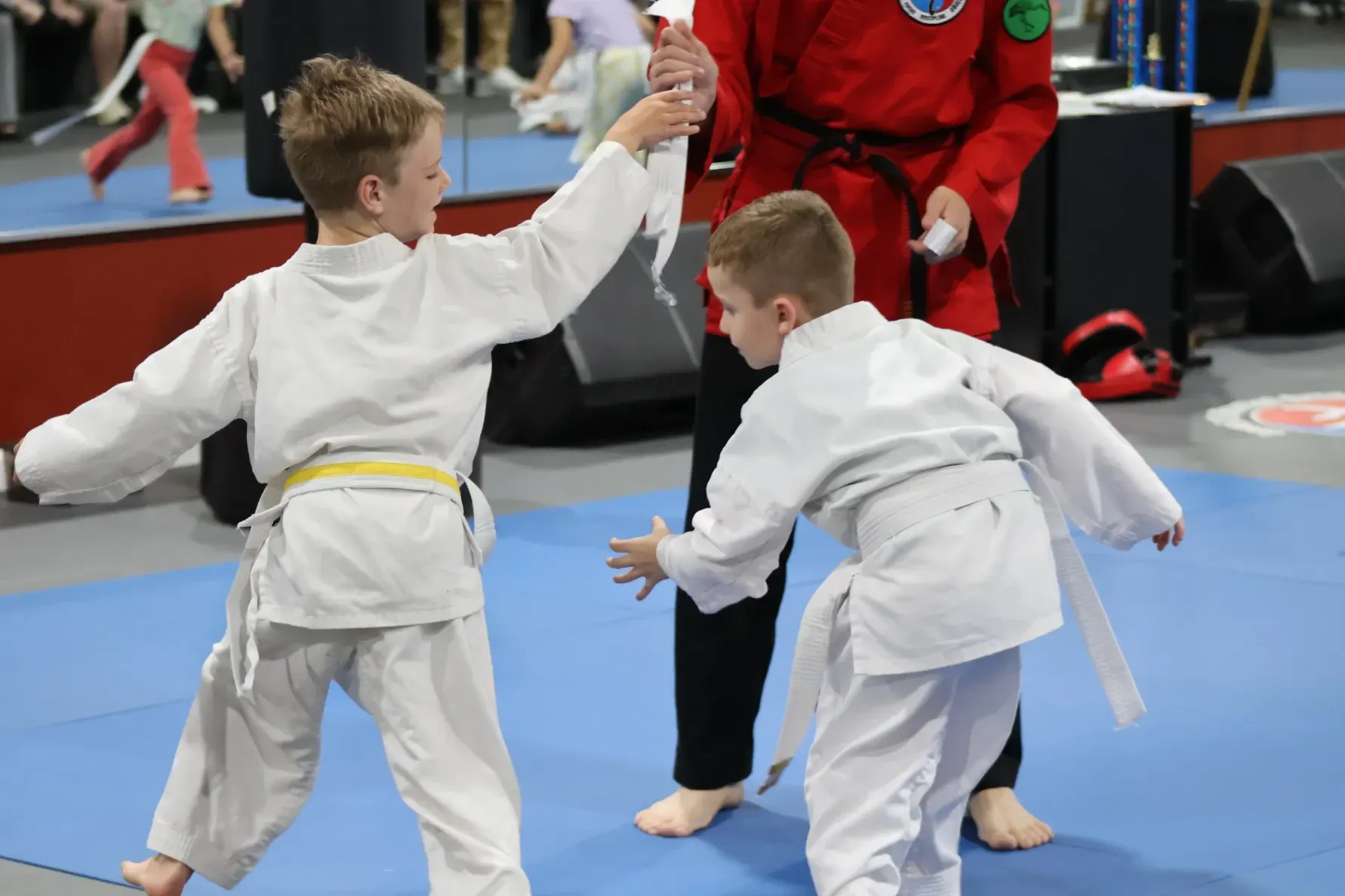 Two children in white karate uniforms spar on a blue mat with a referee in red behind them