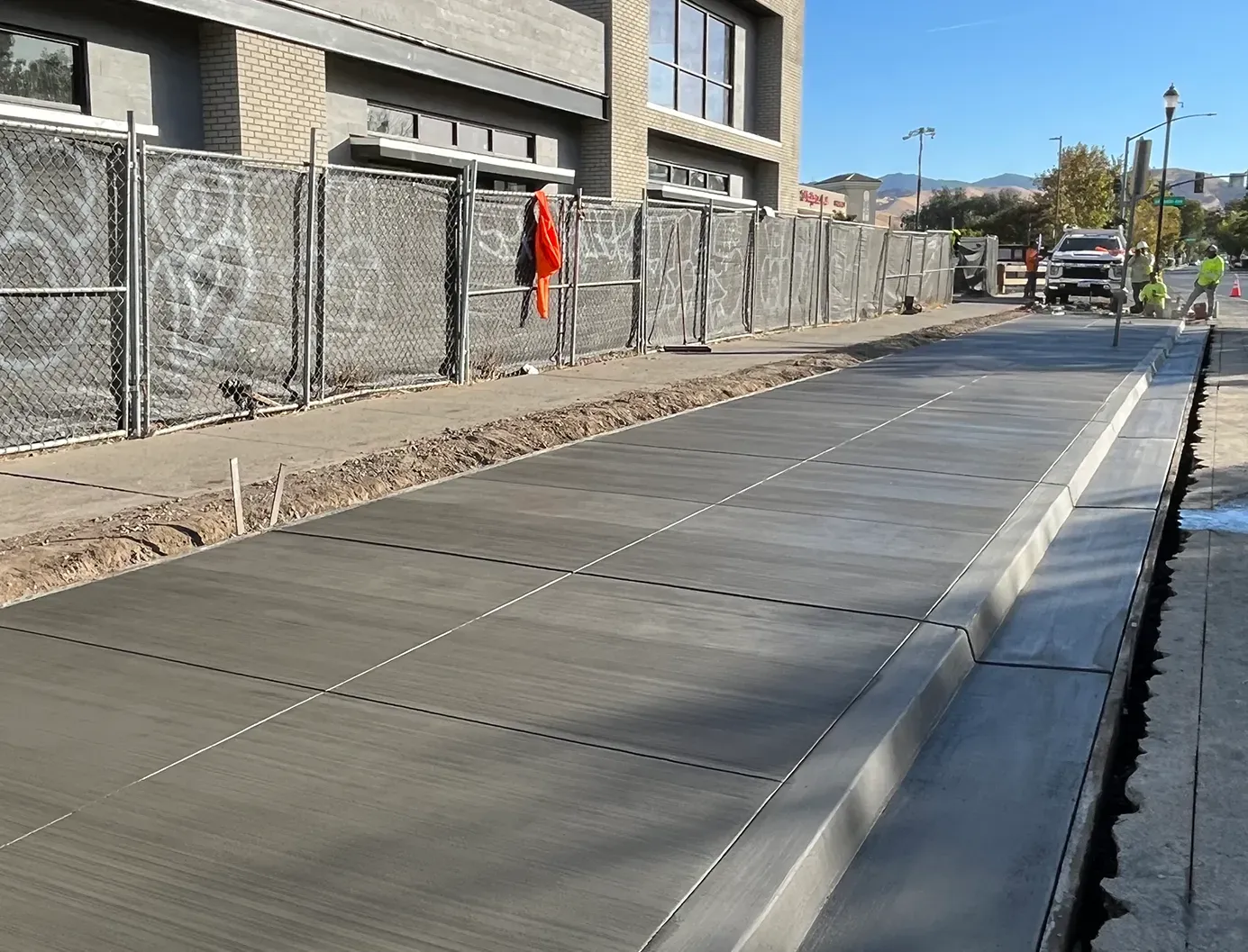 Empty paved street beside a chain-link fence and buildings on a sunny day