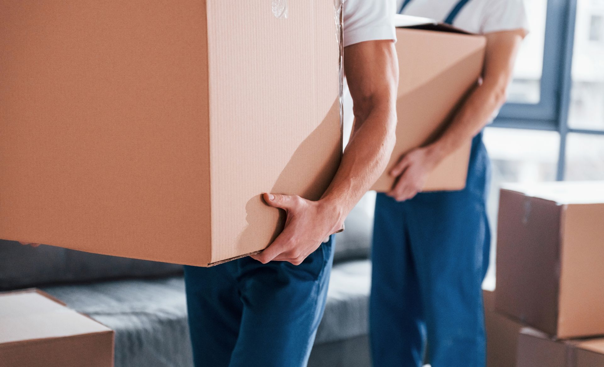 Two men are carrying cardboard boxes in a living room.