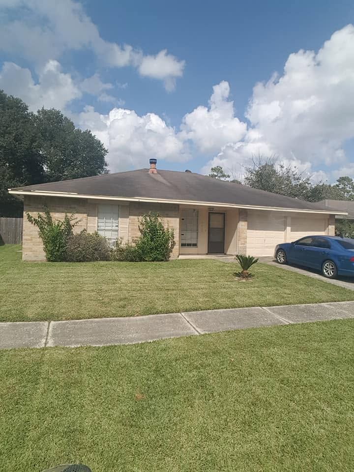 Single-story beige house with a dark roof, blue car, and green lawn under a partly cloudy sky