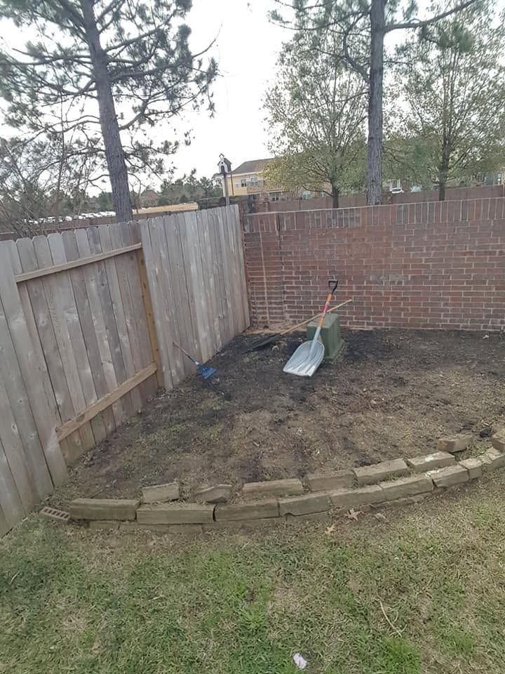Backyard garden bed with shovel, bordered by wooden fence and brick wall, under trees