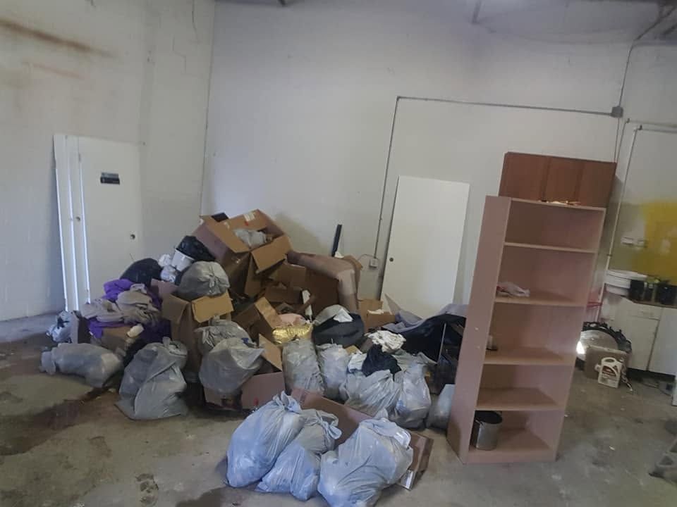 Cluttered room with piled trash bags, cardboard boxes, and a wooden shelf against bare walls.