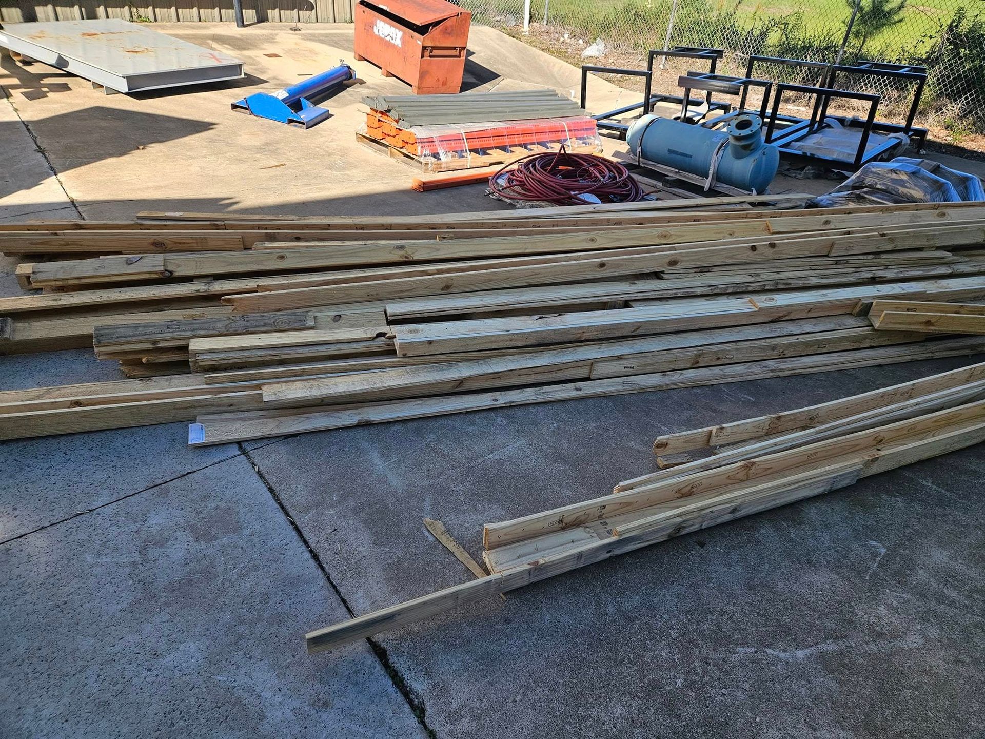 Stacked wooden beams and lumber on a concrete lot with construction tools and equipment in the background