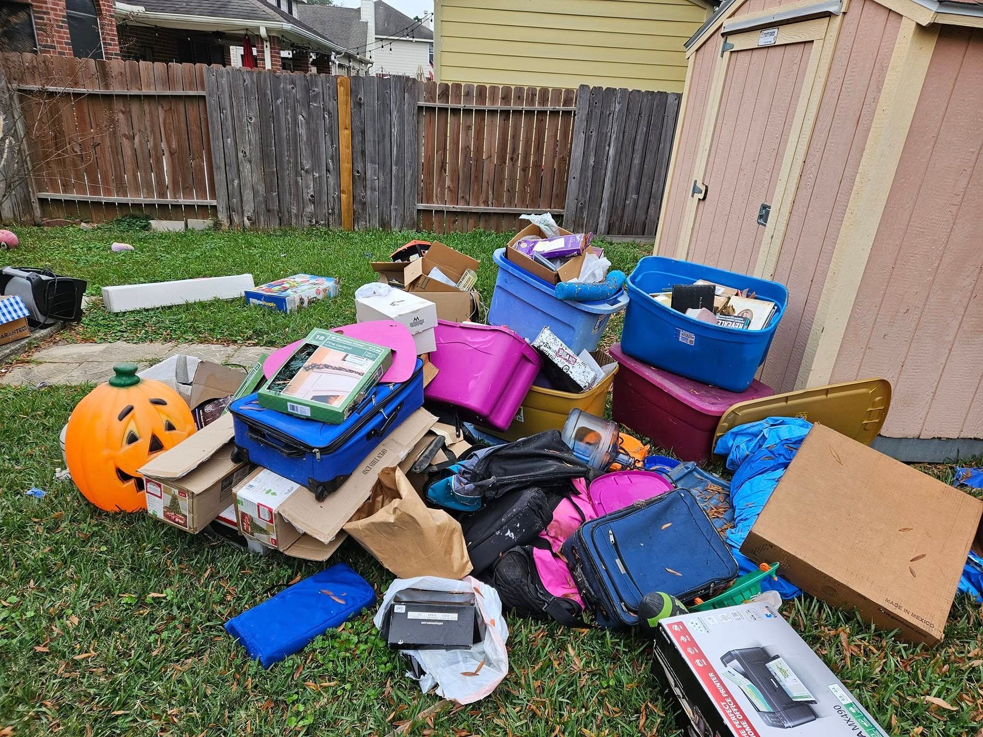 Pile of assorted bags, boxes, and trash scattered on grass beside a wooden fence and shed.