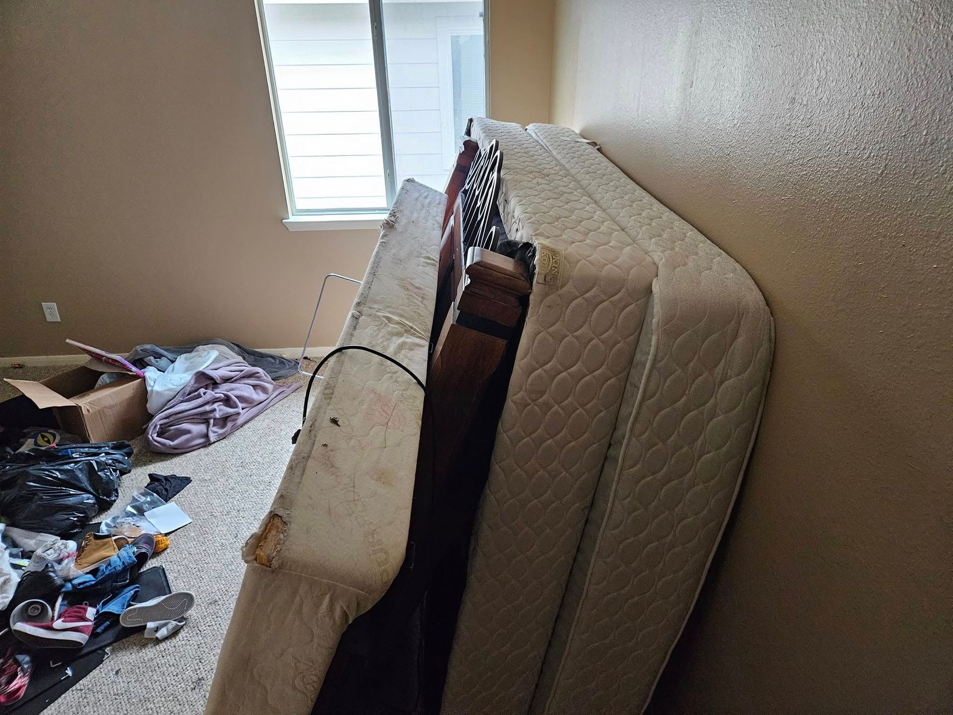 Mattress leaning against a wall in a messy bedroom with clothes and items scattered on the floor.